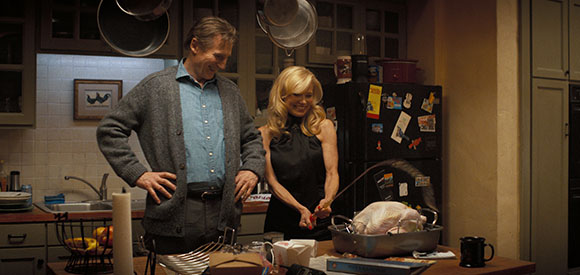 A man in a grey cardigan and a blonde woman in a black dress stand in a kitchen by a roasted turkey.