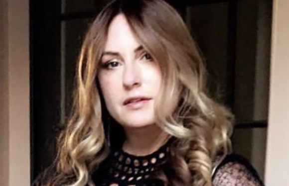 A woman with long, wavy blonde hair and a black lace top looks toward the camera.