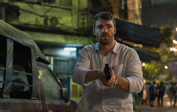 A man in a light blue shirt points a handgun forward in a dimly lit urban street at night.