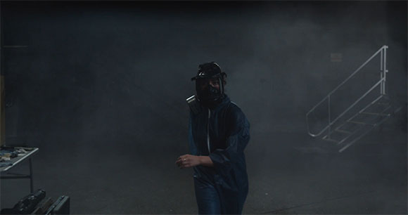 A person wearing a dark protective suit and a full-face mask walks through a dark, smoky room.