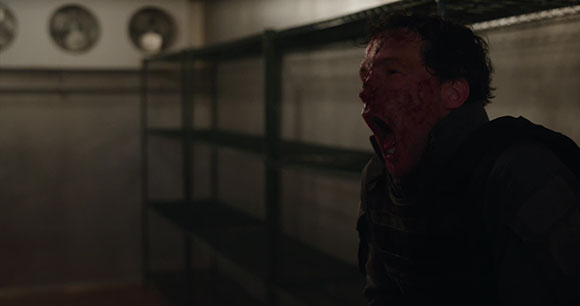 A man with blood on his face screams intensely in a dimly lit room next to metal shelving.