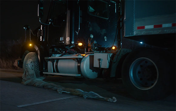 A large cargo net drags along the pavement behind a moving semi-truck at night.