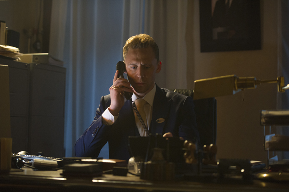 A man in a dark suit sits at a desk and speaks into a black telephone handset.