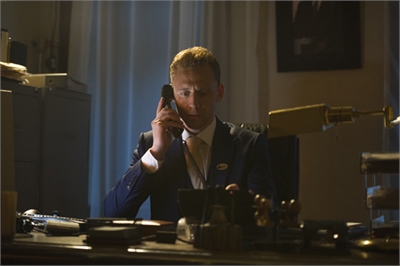 A man in a dark suit and tie speaks into a telephone handset while sitting at a desk in a dimly lit office.