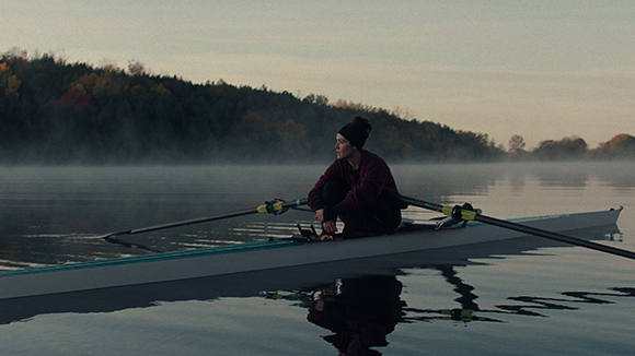 A person in dark clothing sits in a rowing scull on a calm, misty lake at dawn.