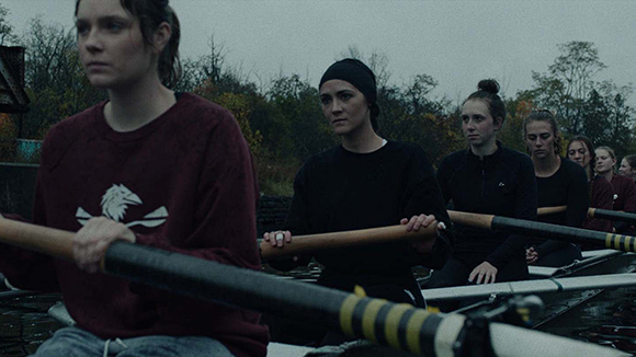 Four young women sit in a rowing shell holding oars on a gray, overcast day near a treeline.