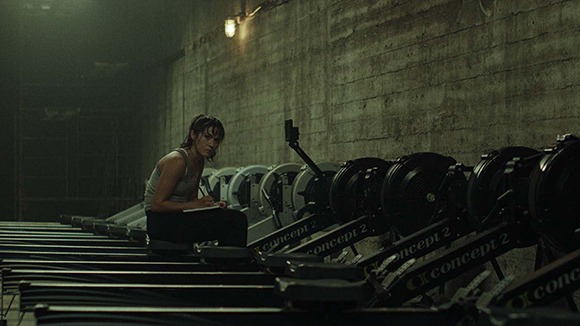 A woman sits on a rowing machine in a dimly lit, concrete industrial gym.