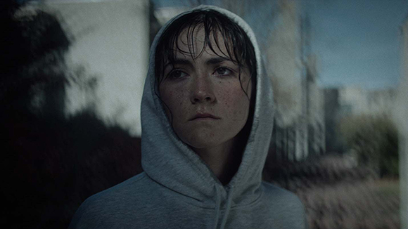 A young woman with freckles and wet hair wearing a grey hooded sweatshirt, looking ahead with a serious expression.