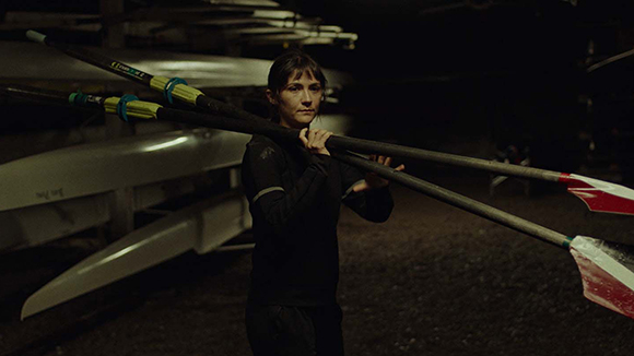 A woman in dark athletic gear carries two long rowing oars over her shoulder in a dimly lit boathouse.