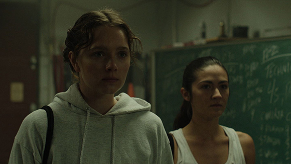 Two young women stand in a dimly lit classroom in front of a green chalkboard.