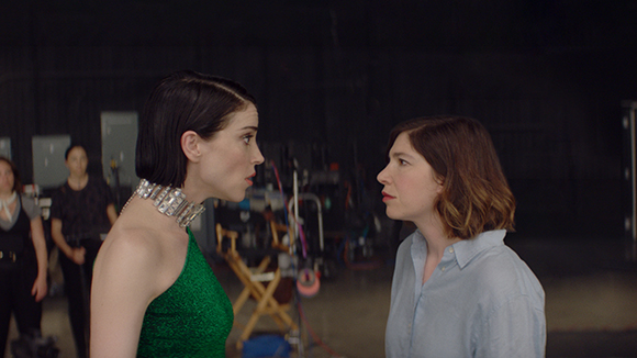 A woman in a green dress and silver choker looks intensely at a woman in a light blue shirt.