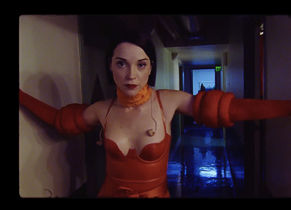 A woman wearing an orange top stands in a hallway with her arms spread wide.