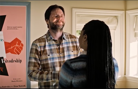 A man in a plaid shirt looks up with a distressed expression while facing a woman with long braids.