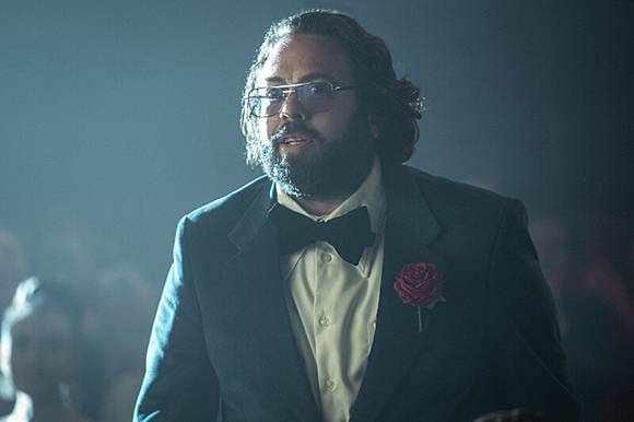 A man with dark hair, glasses, and a beard wearing a black tuxedo and a red rose boutonniere.
