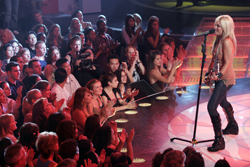A blonde woman in a tan top and dark pants sings into a microphone on a stage in front of a seated audience.