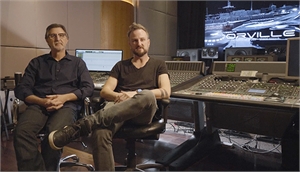 Two men sit in chairs in a recording studio next to a large mixing console.