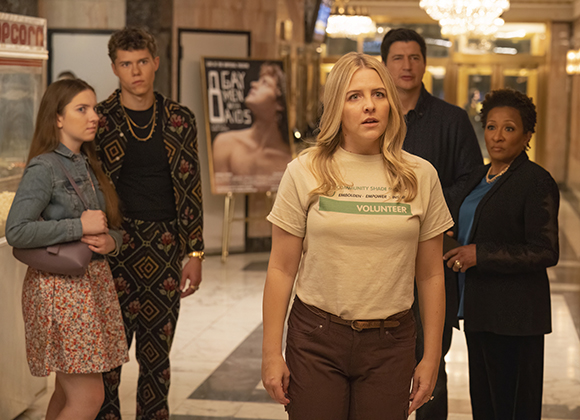 Five people stand in a lobby with surprised expressions, including a woman in a volunteer t-shirt in the foreground.