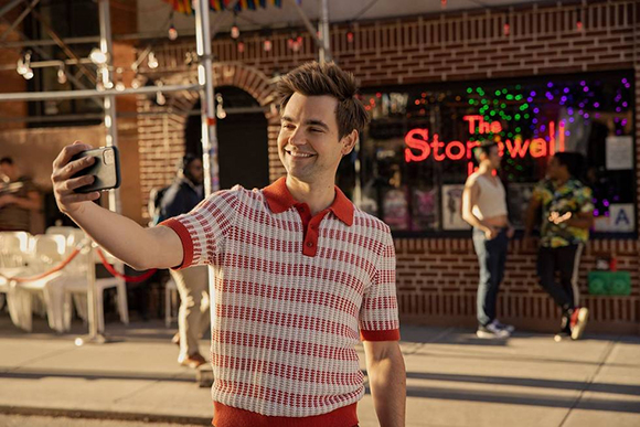 A smiling man in a red and white polo shirt takes a selfie on a city street in front of a brick building.
