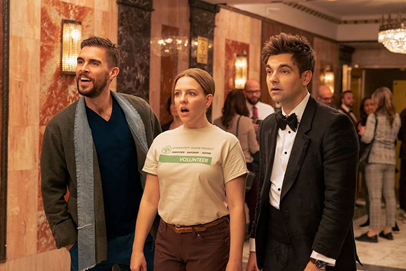 A man in a grey cardigan, a woman in a white volunteer t-shirt, and a man in a tuxedo look on in surprise.