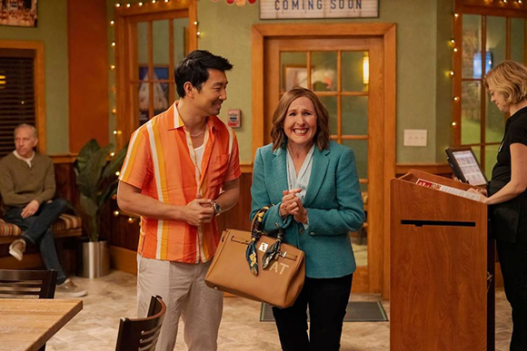 A man in an orange striped shirt and a woman in a teal blazer stand near a wooden podium in a restaurant.