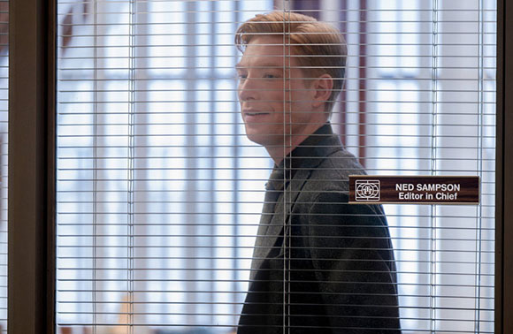 A man in a dark suit looks through horizontal office window blinds.