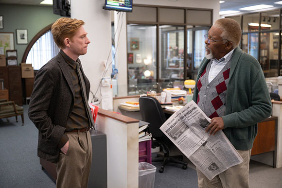 A young man in a brown blazer talks to an older man holding a newspaper in an office.