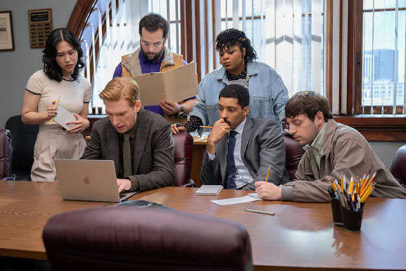 Five diverse professionals gather around a wooden table with a laptop, notebooks, and pens in an office.