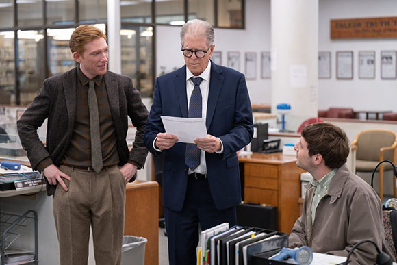A man in a suit reads a paper while two other men look on in an office.