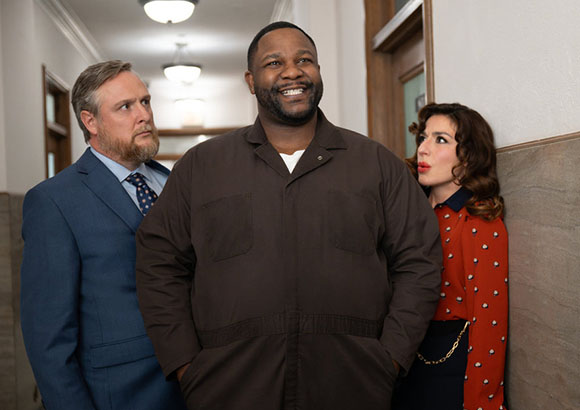 A man in a brown jumpsuit stands between a man in a blue suit and a woman in a red polka dot blouse.