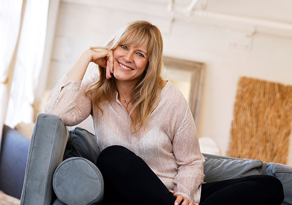 A smiling blonde woman in a light pink blouse sitting on a blue sofa in a bright room.