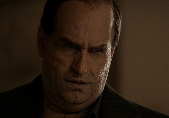 A man with a stern expression and prominent scars on his cheeks looks directly at the camera.