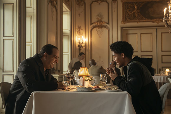 An older man in a dark suit and a younger man in a black hoodie talk at a white-clothed table in a formal dining room.