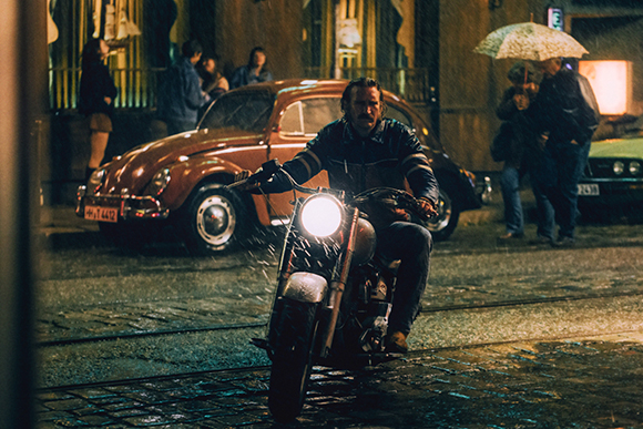 A man on a motorcycle with a bright headlight rides down a wet, cobblestone city street at night.