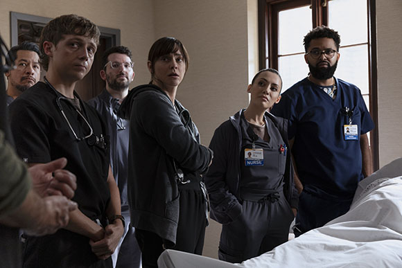 A group of five medical staff members in scrubs looking toward something off-camera in a hospital room.