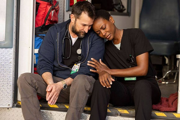 A man and woman in medical scrubs sit close together, leaning their heads against each other looking somber.