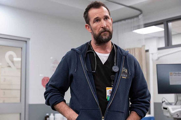 A bearded man in dark blue scrubs and a navy hoodie wearing a stethoscope around his neck.