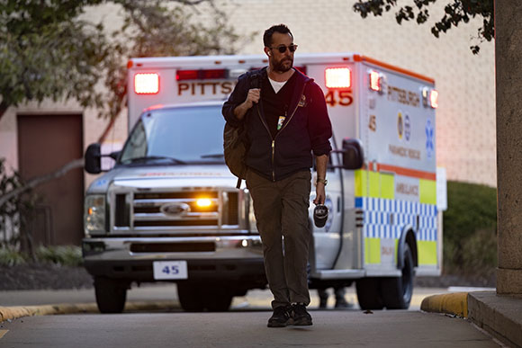 A man in a dark hoodie and sunglasses walks past a white and orange Pittsburgh ambulance.