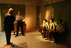 A man in a suit talks to a uniformed officer in a worn room with three people sitting against a wall.