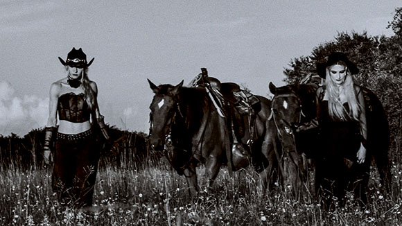 Two women in dark western clothing stand with two horses in a grassy field under a clear sky.