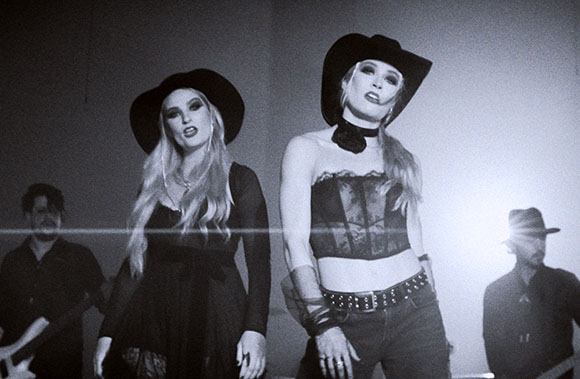 A female singer in a cowboy hat and corset stands center, flanked by band members in a dark setting.