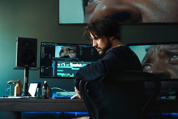 A man sits in a chair facing three computer monitors displaying video editing software and close-up facial shots.