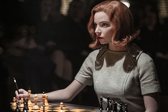 A woman with red hair in a grey dress sits at a chess table, looking intensely at the board.