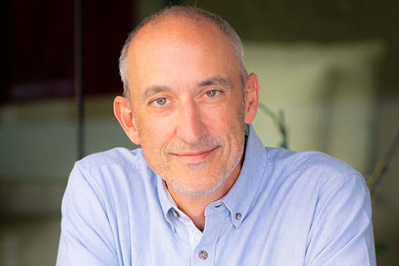 A middle-aged man with short graying hair and a light beard smiling in a light blue shirt.
