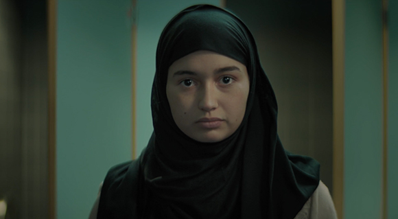 A young woman with a neutral expression wearing a black hijab against a teal background.