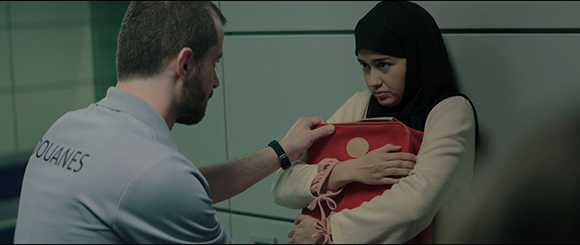 A man in a light blue shirt with 'DOUANES' on the back talks to a woman in a hijab holding a red bag.