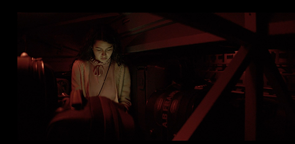 A young woman with dark hair sits in a dark room illuminated by a deep red light.