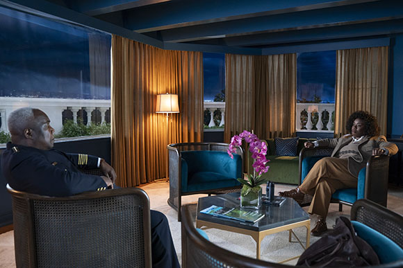 A man in a uniform and a woman in a blazer sit in armchairs in a dimly lit lounge.