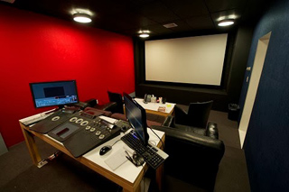 A dark editing studio featuring a red wall, dual monitors, a control console, and a large white projection screen.