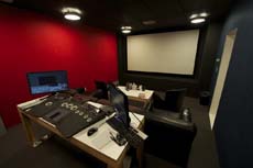 A dark media editing room featuring a red wall, a large projection screen, and a desk with dual monitors and control consoles