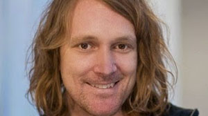 Close-up portrait of a smiling man with long, wavy light brown hair.